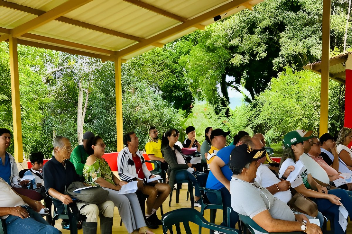 Una asamblea comunitaria reunida en sillas bajo un cobertizo, de espaldas y de perfil.