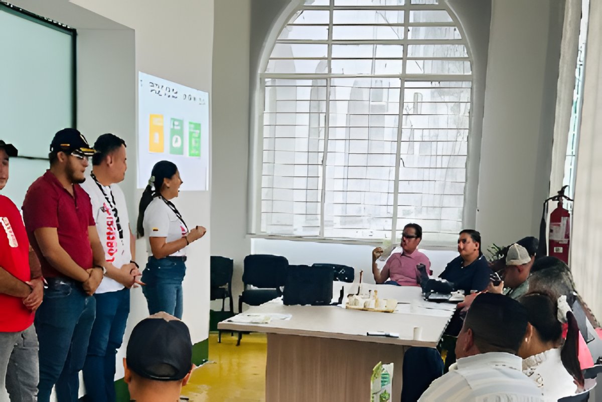 Varias personas de pie exponen ante un grupo sentado alrededor de una mesa, junto a una cartelera.