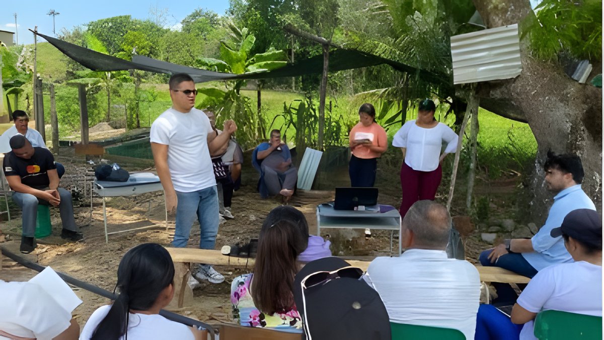 Un grupo sentado en sillas plásticas escucha a un facilitador en una reunión al aire libre, bajo un árbol.