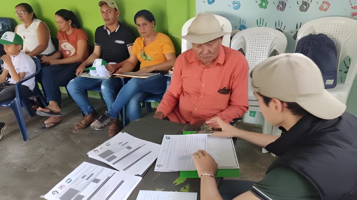 Dos personas revisan formularios sobre una mesa mientras un grupo espera sentado detrás.