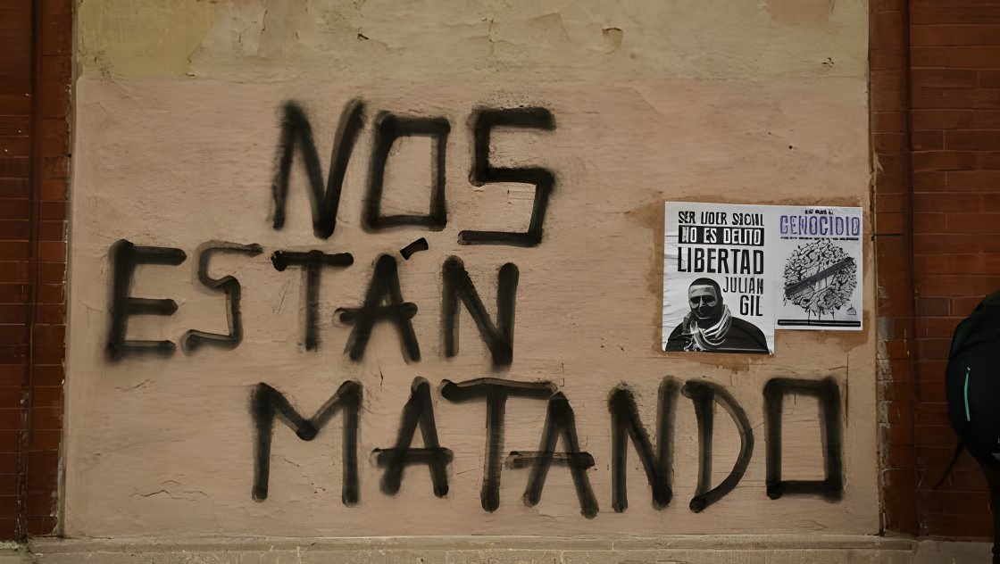 Un muro con la pintada «Nos están matando» junto a carteles pegados.
